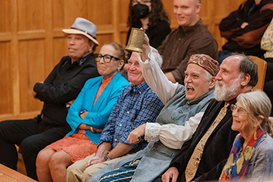 The Host of the Inn wearing a blue renaissance working man's long coat over white puffy shirt and breeches and a cap on his head holds high a tankard as he sits between two men in the front row of the theater, the row and the one behind filled with patrons, all in various modern dress (a guy wearing a tie, another guy where a forora, a woman in bright blue jacke over short orange dress, a guy in a blue plaid shirt, a woman with a colorful scarf.